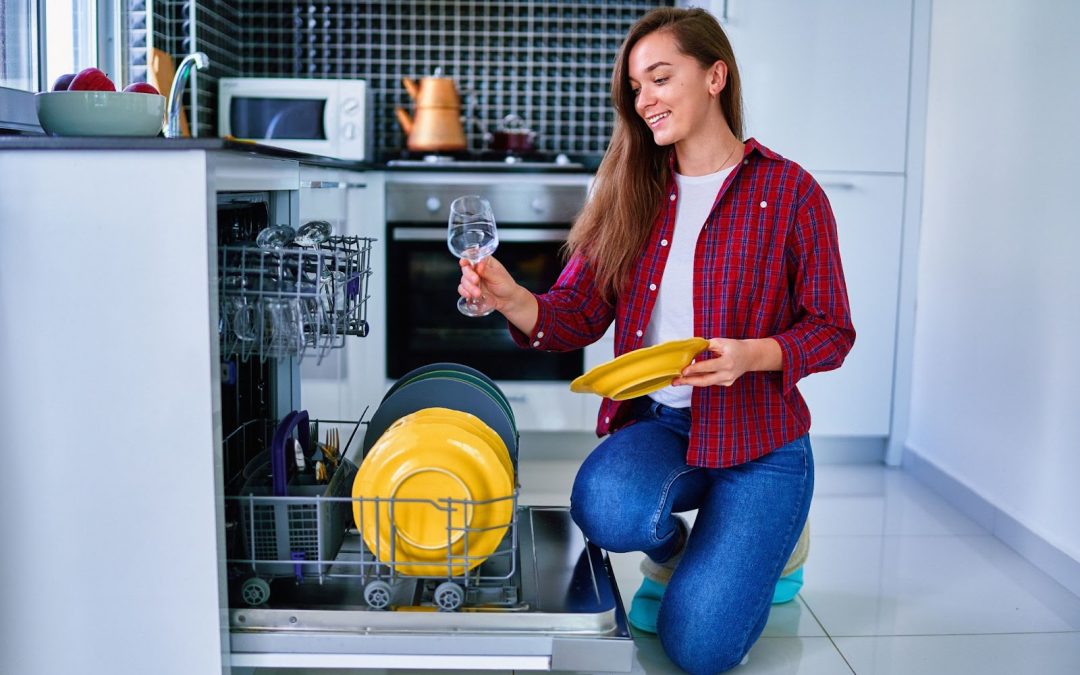 A woman kneels in the kitchen, holding a dish amidst sparkling glassware and a bottle of vinegar for the dishwasher.